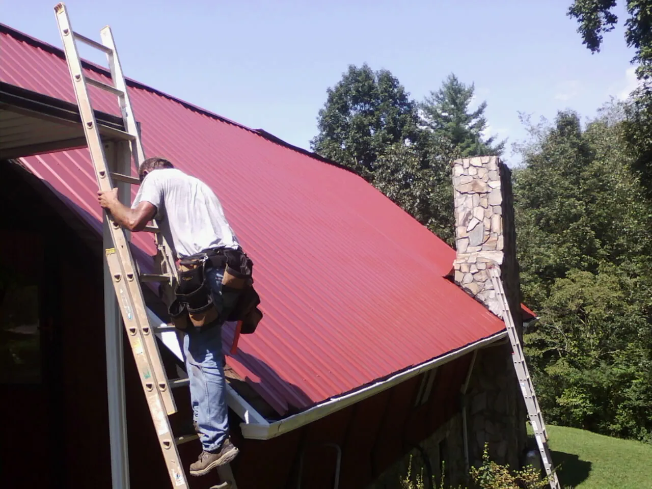 Roofer on a ladder beside a red metal roof and stone chimney — Soffit & Fascia Repair by Roofing Brothers in Pena Blanca